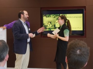 Two people stand in front of a brown wall, smiling and gesturing at each other. Both are holding white pieces of paper with instructions on. In the background a screen reads 'Body Language' with the Reactful logo at the bottom. Reactful CEO Jonathan Friedman is just visible behind them, wearing a purple shirt. In the foreground are the backs of people's heads as they watch the panel.