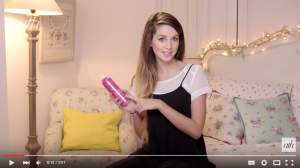 A screenshot from a fishtail braid tutorial by YouTube star Zoella, showing Zoella sitting on a bed and holding a pink can of dry texturising spray.