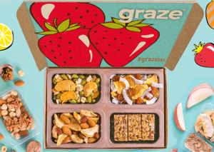 A picture of a Graze summer box with its lid open, with a bold illustration of strawberries inside the lid and four punnets inside. Scattered around it are healthy snack items like pieces of fruit and nuts.