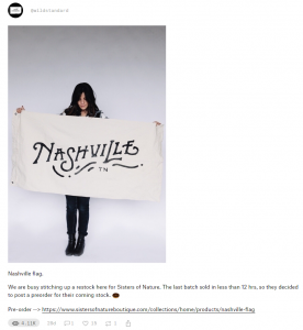 A photograph of a woman holding up a cotton flag which reads "Nashville, TN" in dark, artistic script. Below it the text reads, "Nashville flag. We are busy stitching up a restock here for Sisters of Nature. The last batch sold in less than 12 hrs, so they decided to post a preorder for their coming stock." followed by a doughnut emoji.