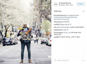 A post from Brompton Bicycle's Instagram showing a man standing in the middle of a street holding a colourful folded Brompton bike. The text reads, "It's weekend time!"