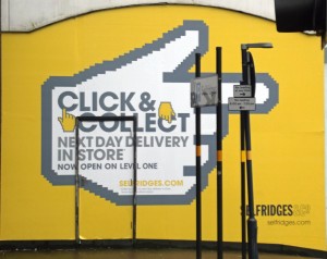 A photograph showing the wall of a Selfridge's, painted yellow with a huge cursor-style hand pointing to the right. Large text reads, Click & Collect, next day delivery in store'.