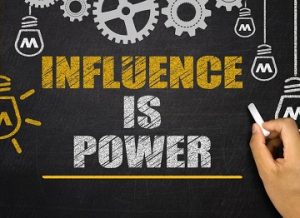 A hand writing on a blackboard with INFLUENCE IS POWER written on it in stylized text, framed by a design of cogs and lightbulbs.